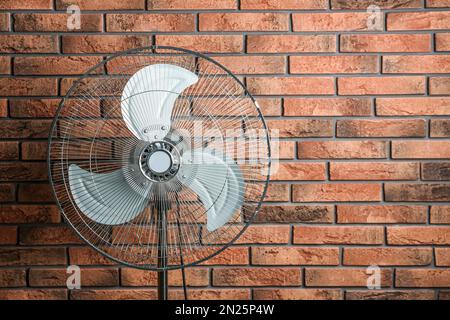 Ventola elettrica vicino alla parete di mattoni, spazio per il testo. Caldo estivo Foto Stock