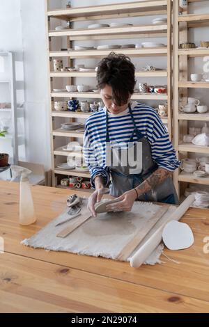 L'artista della ceramica femminile creativa si alza a tavola nel proprio laboratorio e scolpisce il piatto di argilla Foto Stock