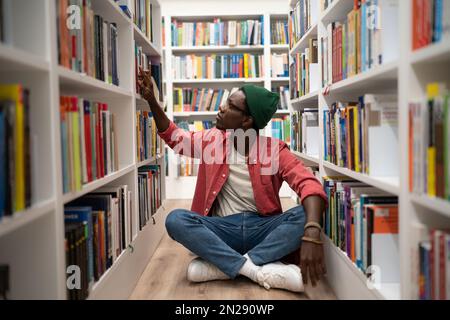 Giovane afro americano ragazzo siede sul pavimento in biblioteca tra librerie, studente alla ricerca di un libro Foto Stock