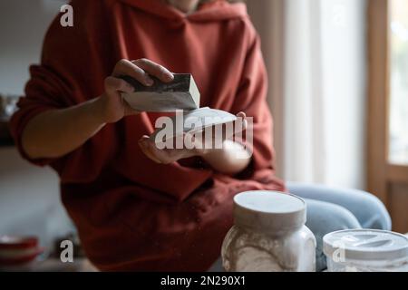 Primo piano mani ceramista donna con piattino e spugna dura per la lucidatura piatti dopo forno di ceramica Foto Stock