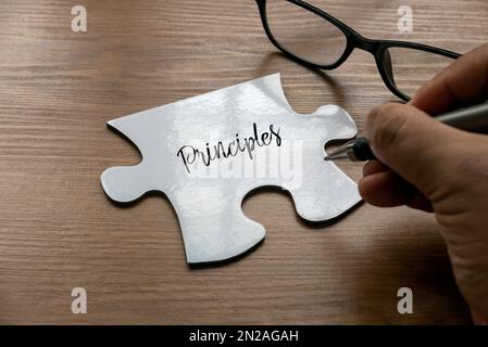 Fuoco selettivo di una parola di scrittura della mano ' principi ' su un pezzo di puzzle bianco con un vetro su sfondo di legno. Foto Stock