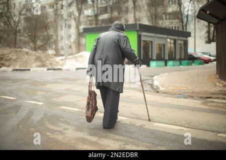 Vecchia donna cammina giù per strada in giacca nera. Pensionato in Russia sulla strada. Nonna con bastone da passeggio. Foto Stock
