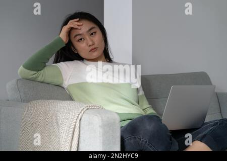 Una donna asiatica che procrastina si siede sul divano con il laptop in grembo e guarda via in modo ingiurioso Foto Stock