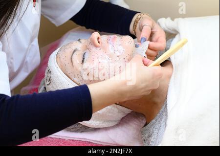 Un estetista rimuove una maschera idratante del foglio sul viso di una donna con una spatola, procedure cosmetologiche. Foto Stock