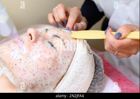 Un estetista rimuove una maschera idratante del foglio sul viso di una donna con una spatola, procedure cosmetologiche. Foto Stock