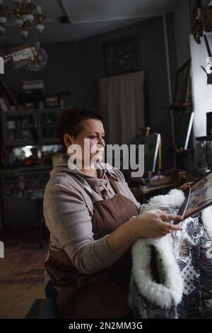 un'artista femminile è seduta su una sedia nello studio guardando un dipinto nella luce dalla finestra. Foto Stock