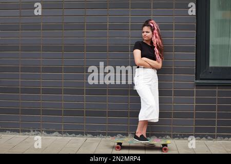 Triste ragazza adolescente con skateboard si trova a tutta lunghezza sulla strada accanto al muro di mattoni grigi. Crisi di adolescenza. Autentico stile di vita ritratto di teena Foto Stock