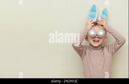 Un ragazzo divertente sotto la veste di un coniglio tiene due giocattoli d'argento opachi, coprendo gli occhi con loro su uno sfondo chiaro con uno spazio per la copia Foto Stock