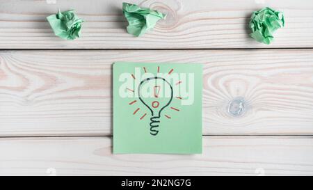 Stendimento piatto di nota adesiva verde con lampadina disegnata con marcatore nero e punto esclamativo rosso all'interno di note quasi stropicciate sul tavolo grigio in legno. Idea Foto Stock