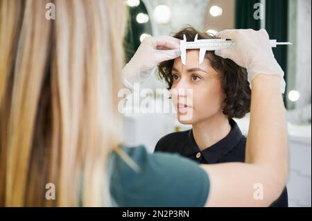 Specialista di bellezza in guanti sterili posizionando il righello sulla fronte della donna durante le misurazioni per la correzione delle sopracciglia. Estetista che utilizza lo strumento di misurazione per la procedura di correzione della fronte in salone di bellezza. Foto Stock