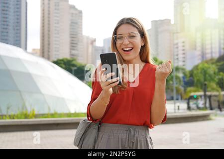 Il brasiliano ha sorpreso la ragazza guardando gioioso il suo smartphone con moderna città sostenibile sullo sfondo Foto Stock