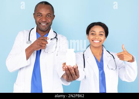 Terapisti maschi e femmine felici in camici bianchi puntando le dita a vaso di farmaci, isolato su sfondo blu Foto Stock