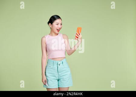 Giovane bella donna asiatica sentirsi felice o sorpresa shock e tenere smartphone su sfondo verde Foto Stock