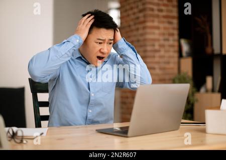 Scioccato uomo d'affari asiatico guardando lo schermo del notebook e toccando la testa, leggendo notizie scioccanti, lavorando da remoto a casa Foto Stock