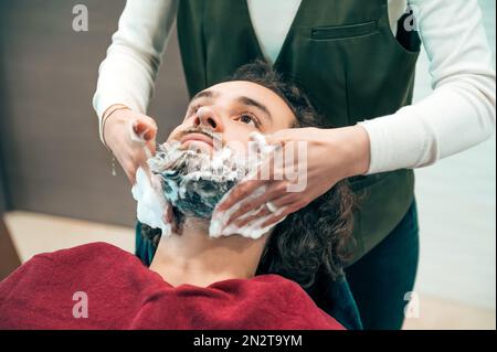 Angolo di raccolto elevato barbiere femmina in uniforme applicazione di sapone sulla barba del cliente mentre si preparano per la procedura di rifilatura in barbiere professionale Foto Stock