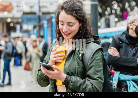 giovane donna latina di etnia argentina, in piedi sulle piattaforme della stazione ferroviaria, utilizzando il telefono per controllare gli orari di partenza del treno e controllare il messaggio Foto Stock