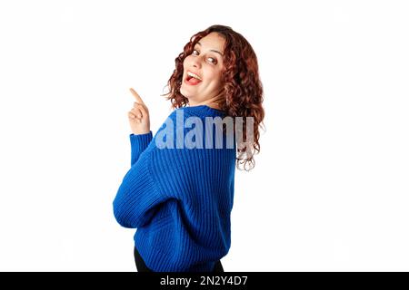 Vista laterale di una donna che indossa un maglione a maglia blu in piedi isolato su sfondo bianco guardando indietro la fotocamera sulla spalla con un sorriso. Confida Foto Stock