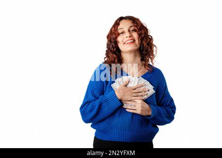 Ritratto di giovane donna che indossa maglia maglione in piedi isolato su sfondo bianco tenendo le banconote vicino al petto, ride e guarda la fotocamera. Foto Stock