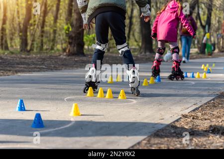 Un gruppo di bambini si diverte imparando la slalom dello skate a rotelle in linea con coni di plastica su strada nel parco cittadino all'aperto nella soleggiata giornata primaverile Foto Stock