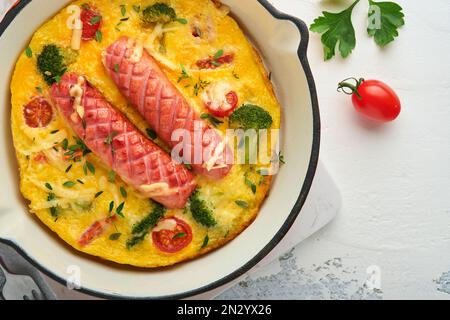 Omelette o frittata con due salsicce, formaggio fuso, pomodori ciliegini, broccoli e prezzemolo su padella rossa o teglia di ferro su fondo bianco di cemento vecchio Foto Stock