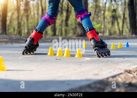 Un gruppo di bambini si diverte imparando la slalom dello skate a rotelle in linea con coni di plastica su strada nel parco cittadino all'aperto nella soleggiata giornata primaverile Foto Stock