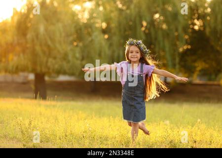 Ragazza carina vestendo corona di fiori all'aperto al tramonto, spazio per il testo. Bambino che passa il tempo in natura Foto Stock