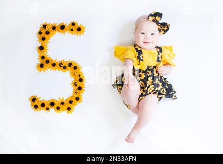 bambina di 5 mesi in un vestito con i fiori. Baby Milestone cinque mesi. Foto Stock