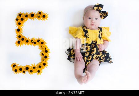 bambina di 5 mesi in un vestito con i fiori. Baby Milestone cinque mesi. Foto Stock