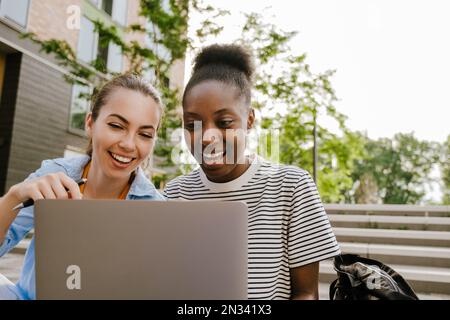 Giovani donne multirazziali che fanno i compiti con il computer portatile mentre si siedono sulle scale all'aperto Foto Stock