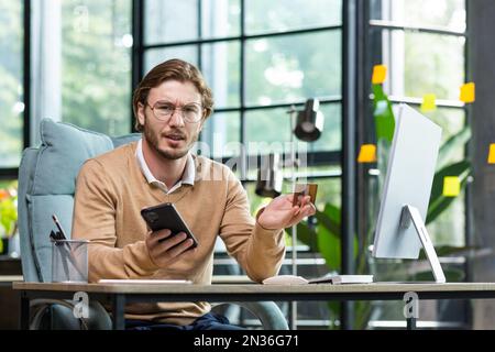 Problema con una carta di credito. Giovane uomo d'affari confuso, libero professionista seduto in ufficio e al tavolo e utilizzando il telefono e la carta di credito. Guarda insoddisfatto la telecamera, agita le mani. Foto Stock
