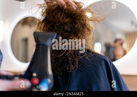 Anonimo stylist che asciuga i capelli ricci del cliente femminile nel salone di bellezza mentre si prepara per il taglio dei capelli Foto Stock