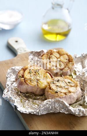 Bulbi all'aglio arrostiti con erbe e olio d'oliva in foglio di stagno su un tagliere Foto Stock