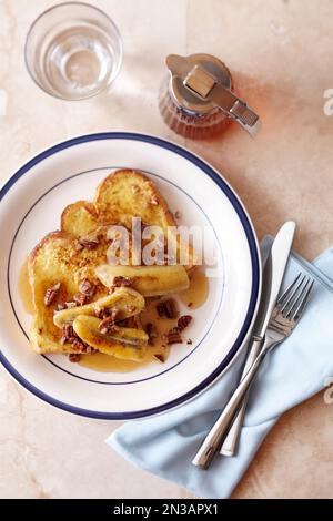 Toast alla francese con banana arrostita, noci pecan e sciroppo d'acero su un piatto bianco con bordo blu Foto Stock