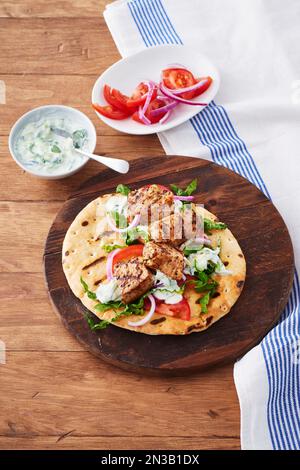 Souvlaki di maiale su una pita grigliata su un tagliere di legno con contorni di tzatziki e pomodori tritati e cipolle a fette Foto Stock