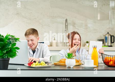 Felici i bambini, i bambini che fanno colazione a casa e utilizzano i dispositivi digitali. i bambini giocano e mangiano. Foto Stock