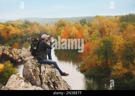 Escursionista con smartphone seduto sulla ripida scogliera vicino al fiume di montagna Foto Stock