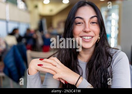 Giovane donna positiva con lunghi capelli scuri in camicia informale sorridente e guardando la macchina fotografica mentre si siede in un bar Foto Stock