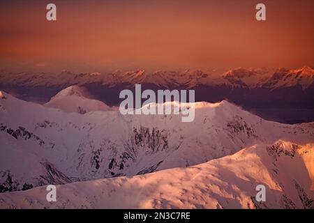 Venti forti soffiano la neve attraverso le creste scoscese e le vette delle South Chilkat Mountains illuminando intensi colori arancioni di un tramonto invernale. Il... Foto Stock