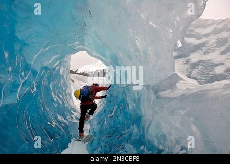 Vista da dietro di un escursionista, dotato di ramponi e attrezzature di emergenza, che scorre attraverso un tunnel di ghiaccio blu formato nel Glac di Mendenhall... Foto Stock