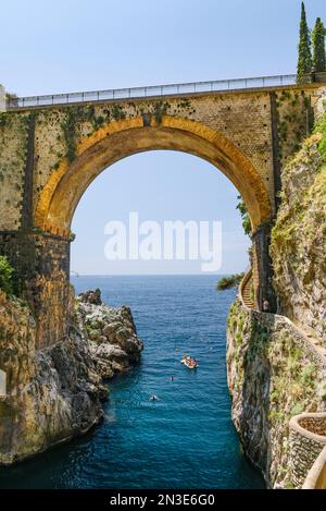 Persone che nuotano e navigano sotto un vecchio ponte stradale attraverso la baia di Furore; Furore, Salerno, Italia Foto Stock
