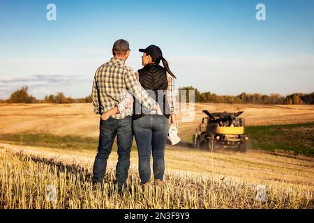 Vista da dietro di un marito e di una moglie in piedi in un campo che trascorrono un po' di tempo insieme mentre guardano una mietitrebbia finire la loro caduta... Foto Stock