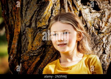 Giovane ragazza che gioca vicino a un albero e si ferma per un momento in un parco cittadino in un caldo pomeriggio autunnale; St. Albert, Alberta, Canada Foto Stock