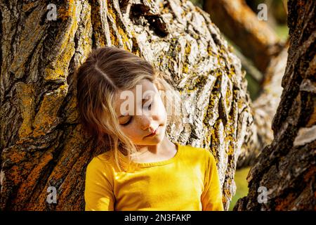 Giovane ragazza che gioca vicino a un albero e si ferma per un momento in un parco cittadino in un caldo pomeriggio autunnale; St. Albert, Alberta, Canada Foto Stock