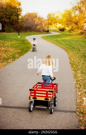 Giovane ragazza che tira il suo carro lungo un sentiero in un parco cittadino durante la stagione autunnale con suo fratello in bicicletta verso di lei Foto Stock