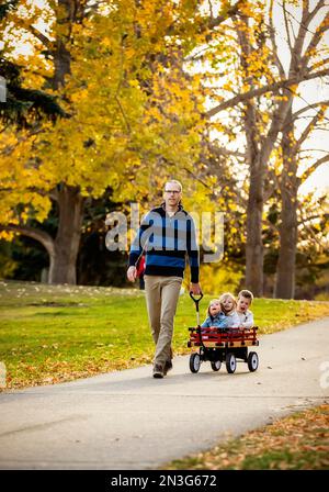 Un padre che tira i suoi figli in un carro lungo un fiume in un parco cittadino con un ponte sullo sfondo durante la stagione autunnale e il suo bambino gir... Foto Stock