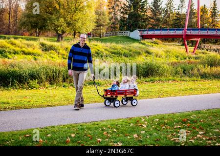 Un padre che tira i suoi figli in un carro lungo un fiume in un parco cittadino con un ponte sullo sfondo durante la stagione autunnale e il suo bambino gir... Foto Stock