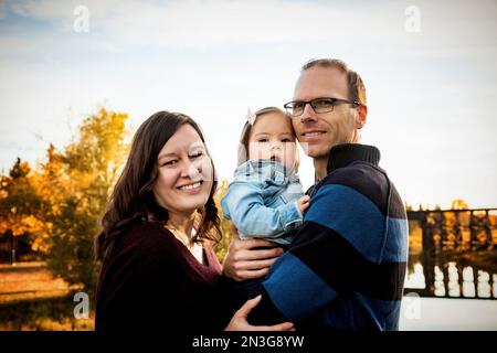 Ritratto di famiglia in piedi su un ponte sopra un fiume in un parco cittadino durante il mare d'autunno e la loro bambina ha la sindrome di Down Foto Stock