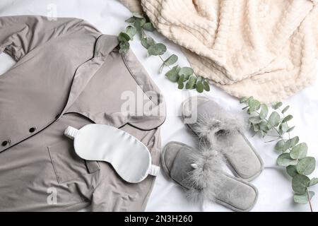 Composizione piatta con soffici pantofole e pigiami su copriletto bianco. Comodo outfit per la casa Foto Stock