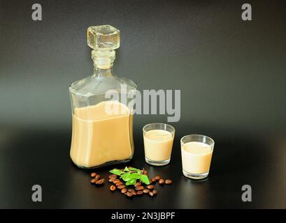 Liquore alla crema su sfondo nero, bottiglia e due bicchieri con alcol fatto in casa, foglie di menta e chicchi di caffè arrostiti nelle vicinanze. Primo piano. Foto Stock