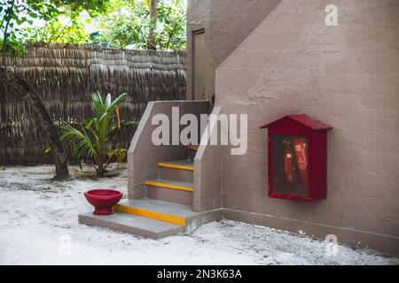 Una vista di una casa di vacanza tropicale entrata con un pavimento di sabbia, recinzione di canne la residenza, alcuni alberi, uno strumento antincendio, una ciotola per il lavaggio dei piedi, Foto Stock